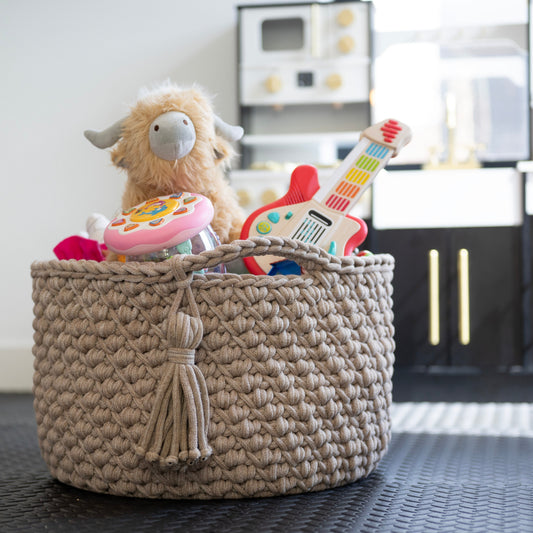 A round woven storage basket filled with toys, including a plush sheep and a toy guitar, set on a dark textured floor in a child's playroom with a toy kitchen in the background.