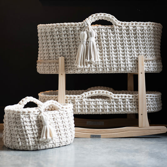 A set of cream woven baby baskets with tassel handles, including a large basket displayed on a wooden rocking cradle and a smaller matching basket in front, set against a dark background.