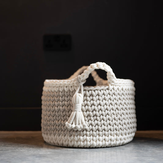A cream-colored crocheted basket with two loop handles and a hanging tassel, set against a dark charcoal wall and a gray stone floor.