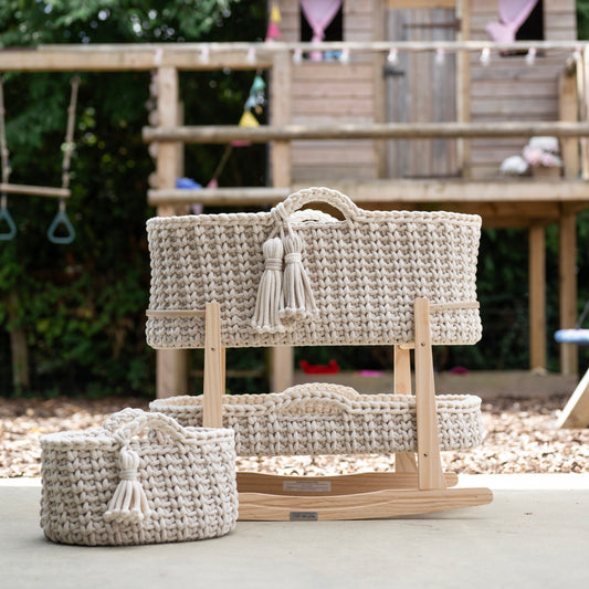 A woven natural-fiber baby bassinet set displayed on a wooden rocking base in an outdoor backyard setting with a wooden playhouse and trees in the background.