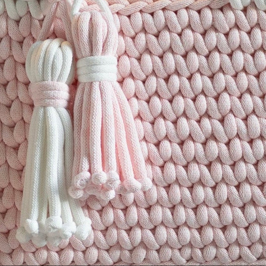 A pink and white crocheted baby bassinet set with wooden rockers, shown with a matching bassinet on the floor against a dark paneled wall with a warm hanging light.
