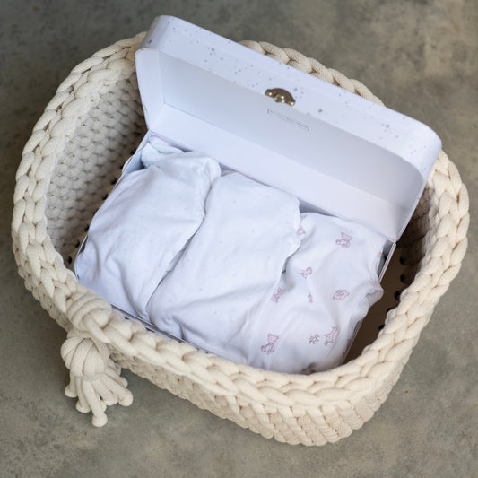 A white gift box with a small gold elephant logo sits open inside a cream woven basket, revealing folded white baby clothes with tiny pink floral prints, set on a gray concrete floor.