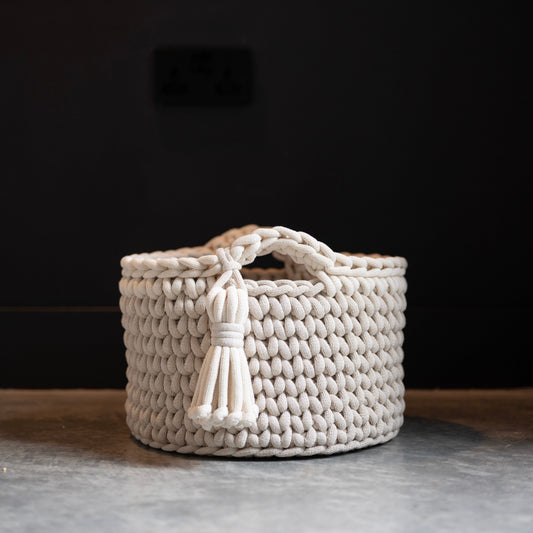 A round, cream-colored woven basket with braided handles and a hanging tassel, set on a gray stone surface against a dark, moody background.