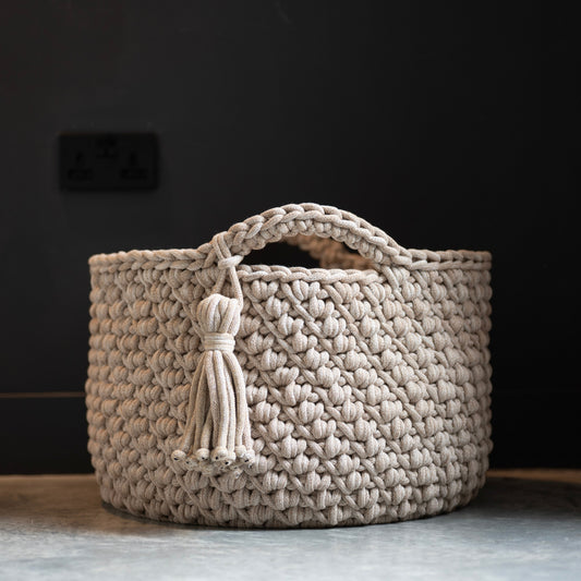 A round, handwoven basket with a top handle and a decorative tassel, shown on a gray floor against a dark background.