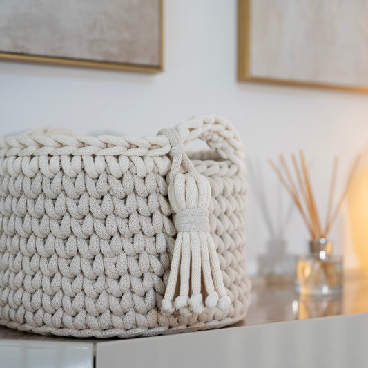 A cream-colored chunky knit storage basket with a tassel handle, styled on a light-colored cabinet next to a diffuser and a ceramic vase.