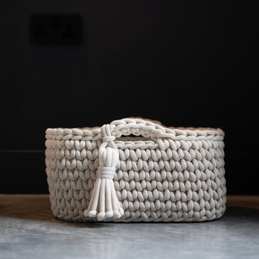 A small woven basket in a light cream or off-white color, featuring a top handle and a decorative tassel, set against a dark background.